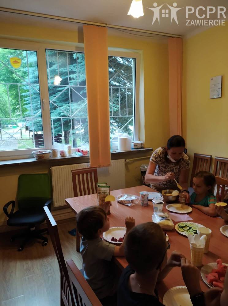 Zdjęcie: Grupa dzieci siedzi przy stole, na którym w pojemnikach poustawiane są pokrojone produkty spożywcze, a kobieta nakłada na talerzyk